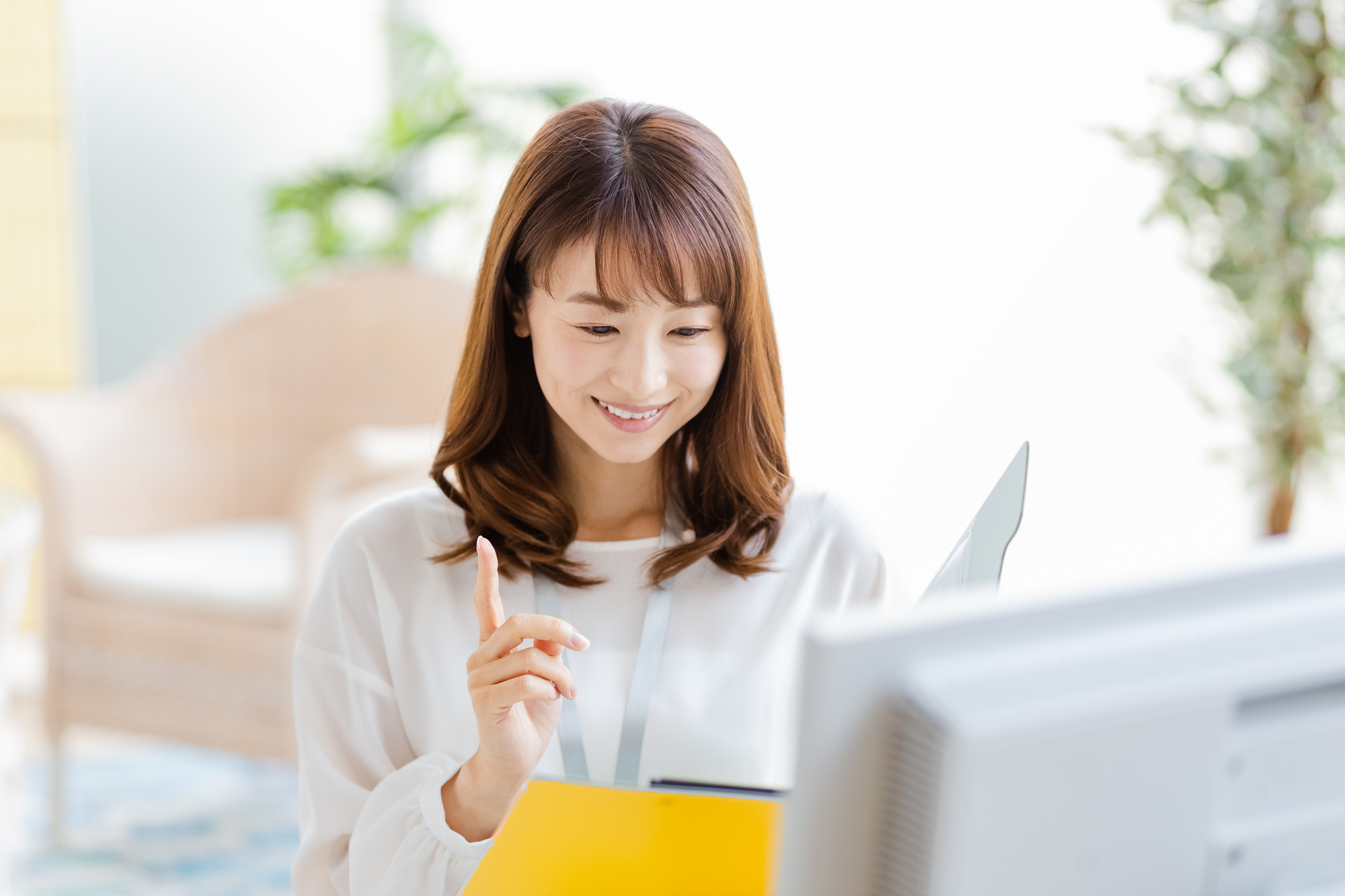 パソコンの前に座って資料を見ながら人さし指を立てている笑顔の女性