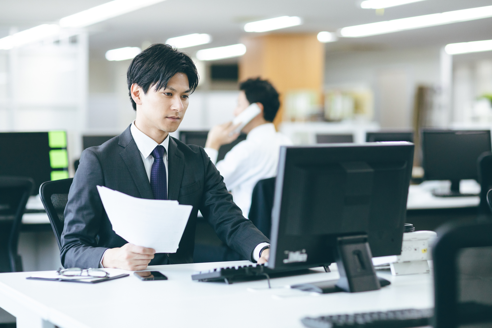 右手に紙を持ち、パソコンを操作するスーツの男性