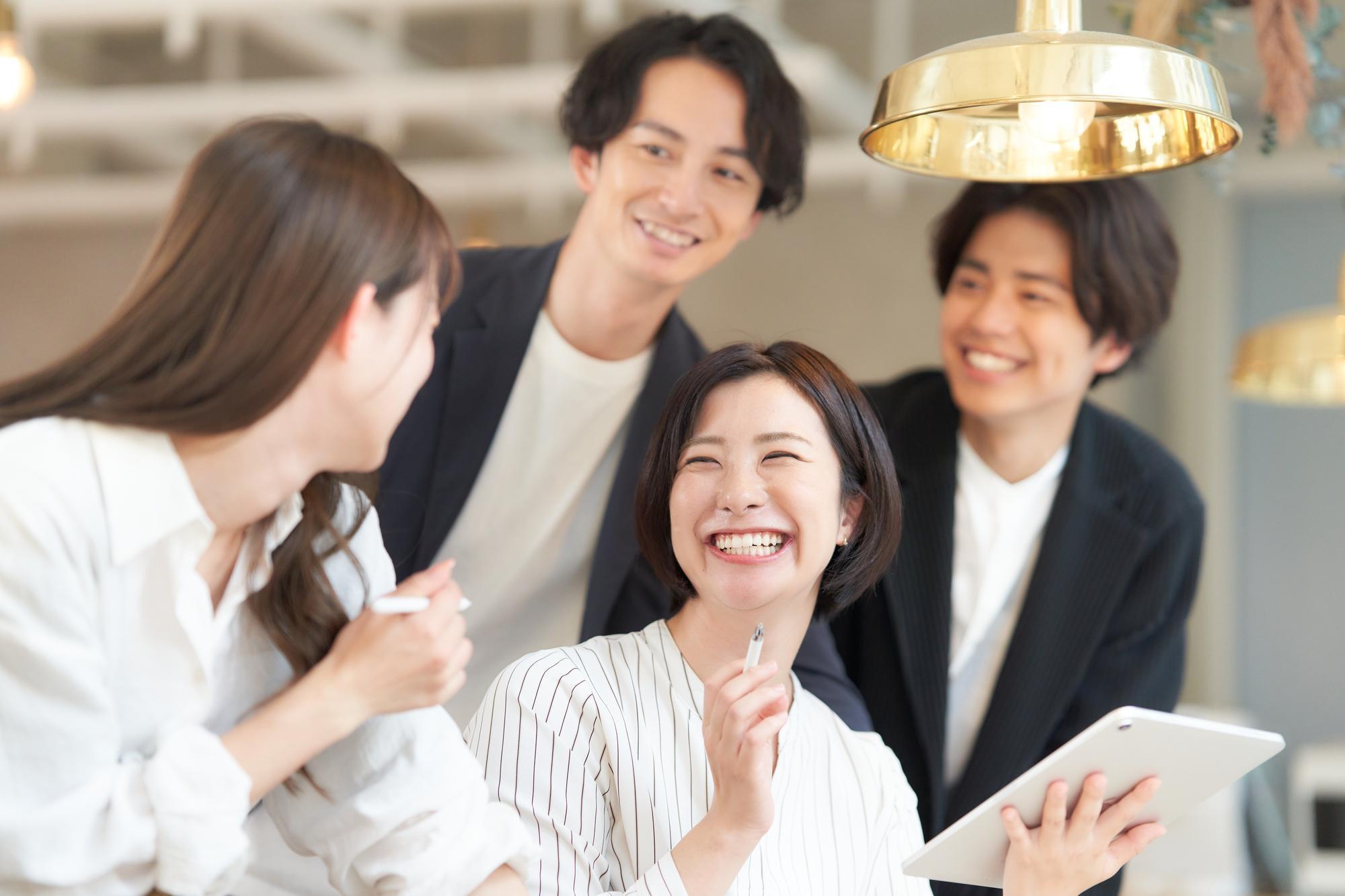 左手にタブレット、右手にペンを持つ笑顔の女性の周りに集まる笑顔の男女