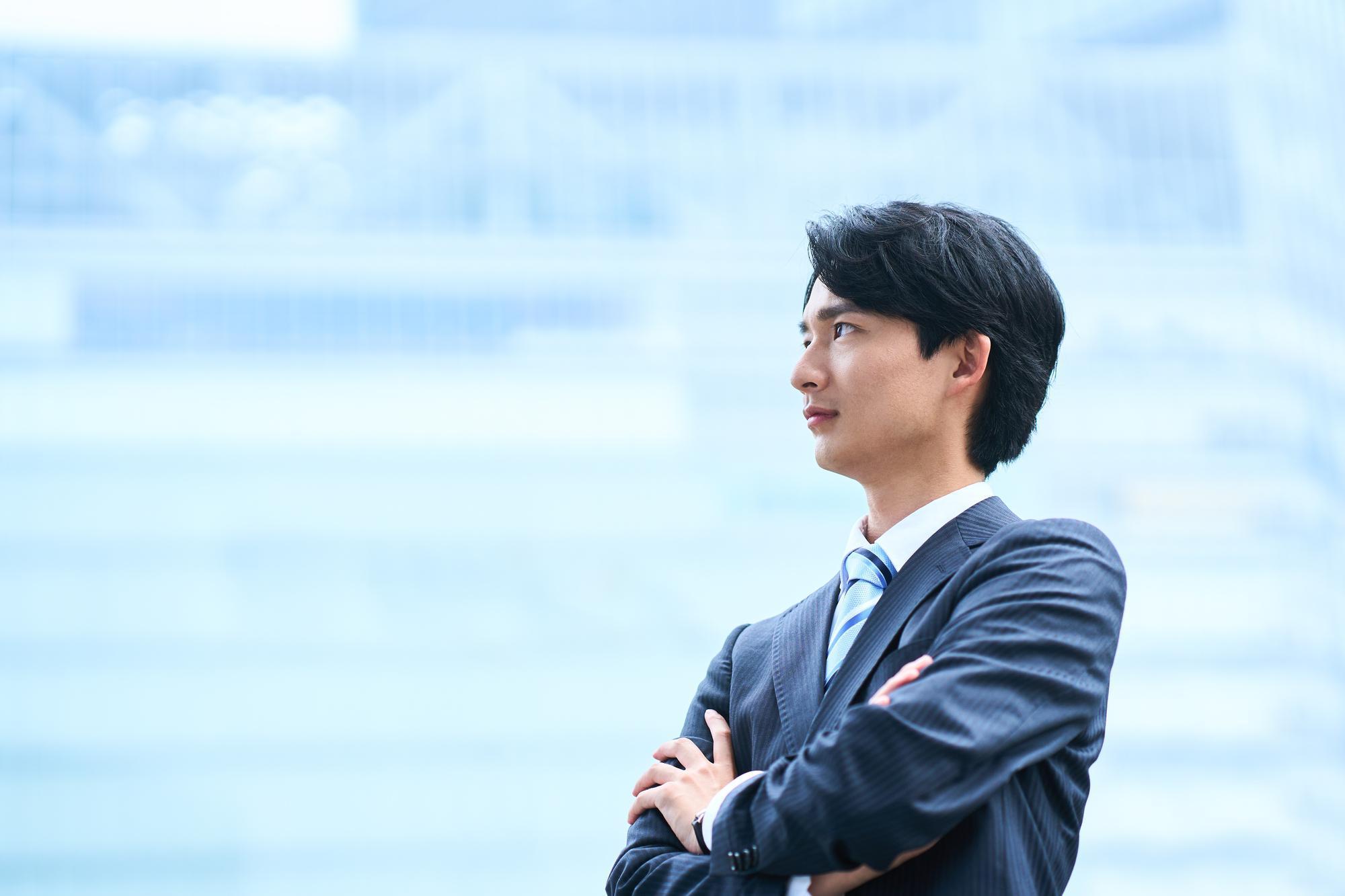 屋外にて、両腕を前に組み、真剣な表情で斜め上を見るスーツ姿の男性