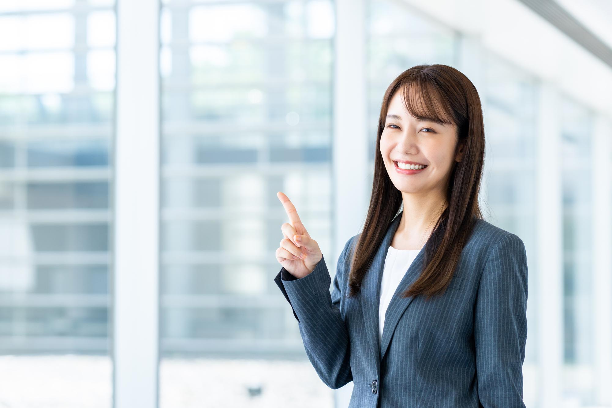 人さし指を立て、カメラ目線で笑顔を向けるスーツ姿の女性