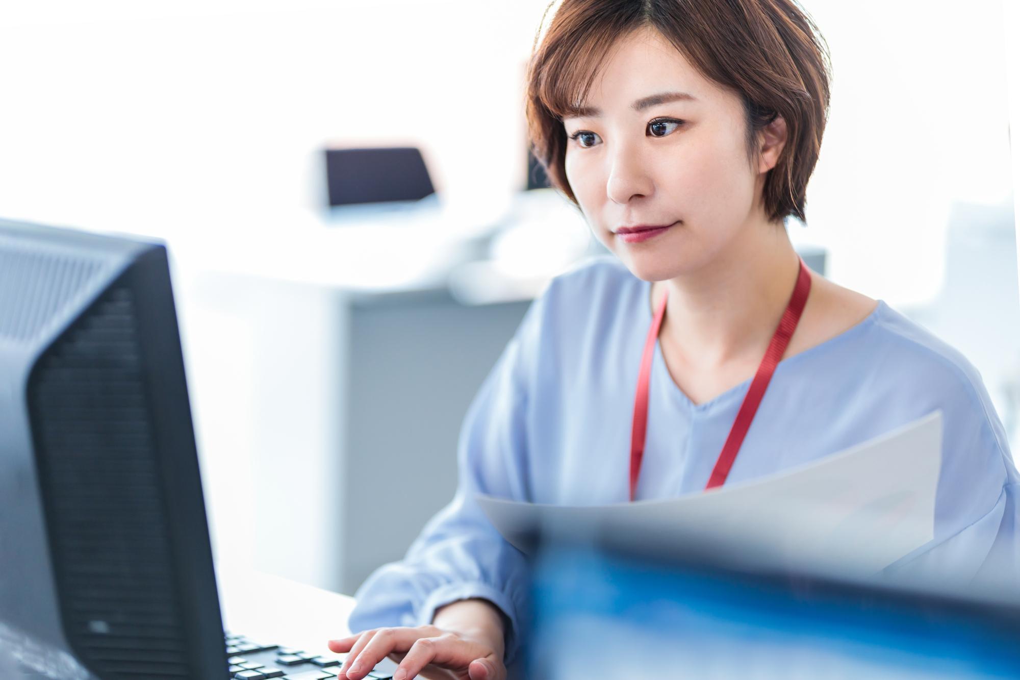 左手に紙を持ち、右手でパソコンを操作する女性