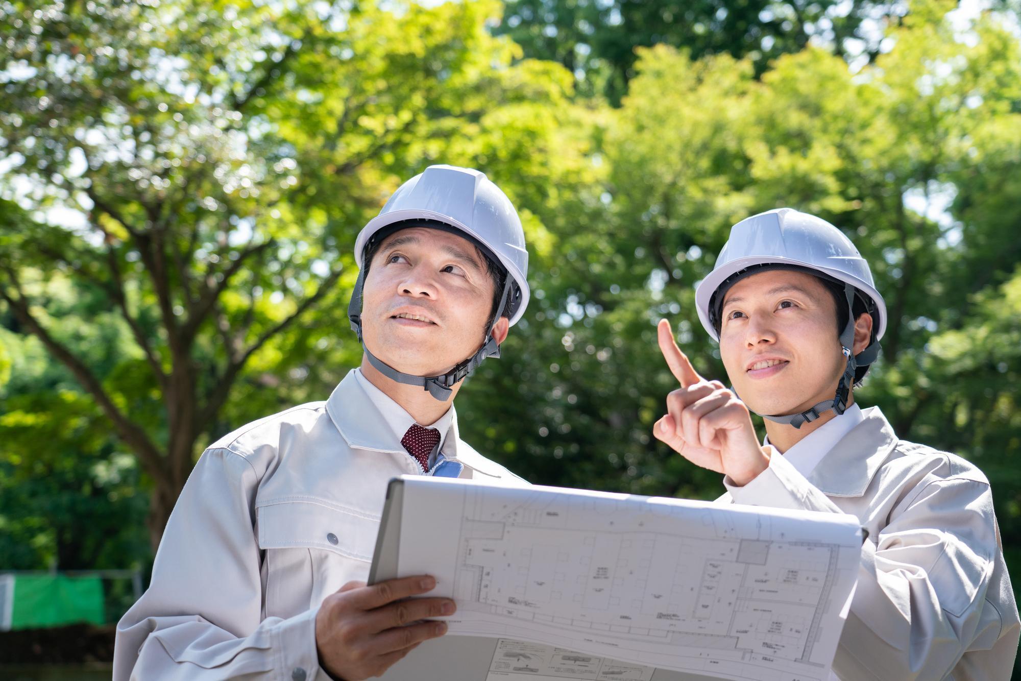図面を持ち、上を見ている、ヘルメットをかぶった作業着姿の2人の男性