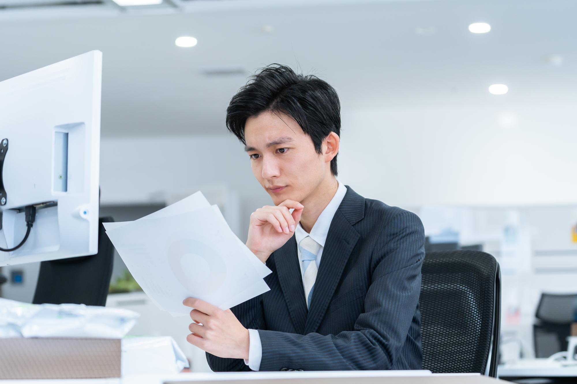 右手を顎に当てて左手に持った紙を見つめ、考えている様子のスーツの男性