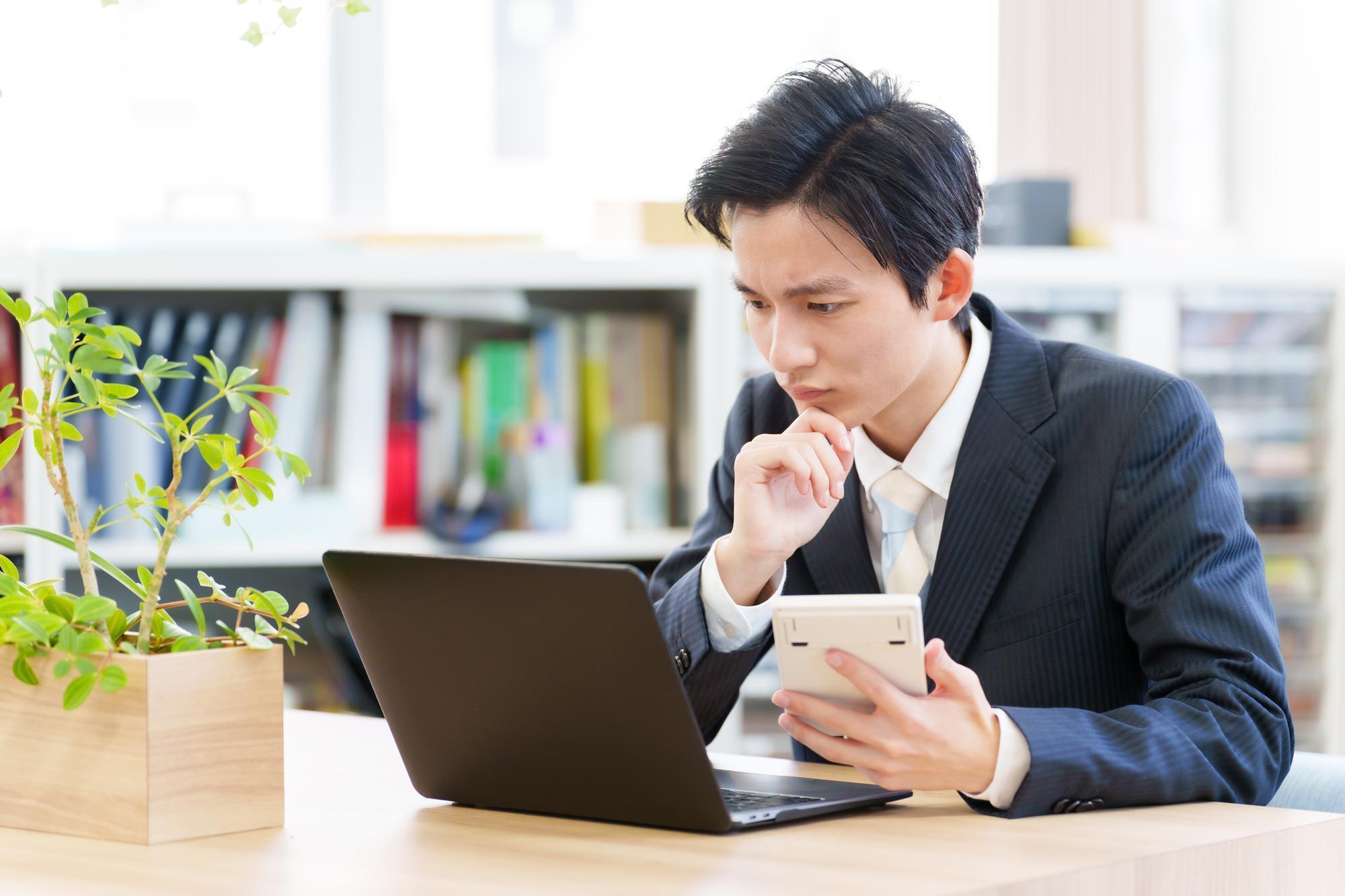 右手を顎に当て、左手に電卓を持ち、真剣な表情でパソコンを見るスーツの男性