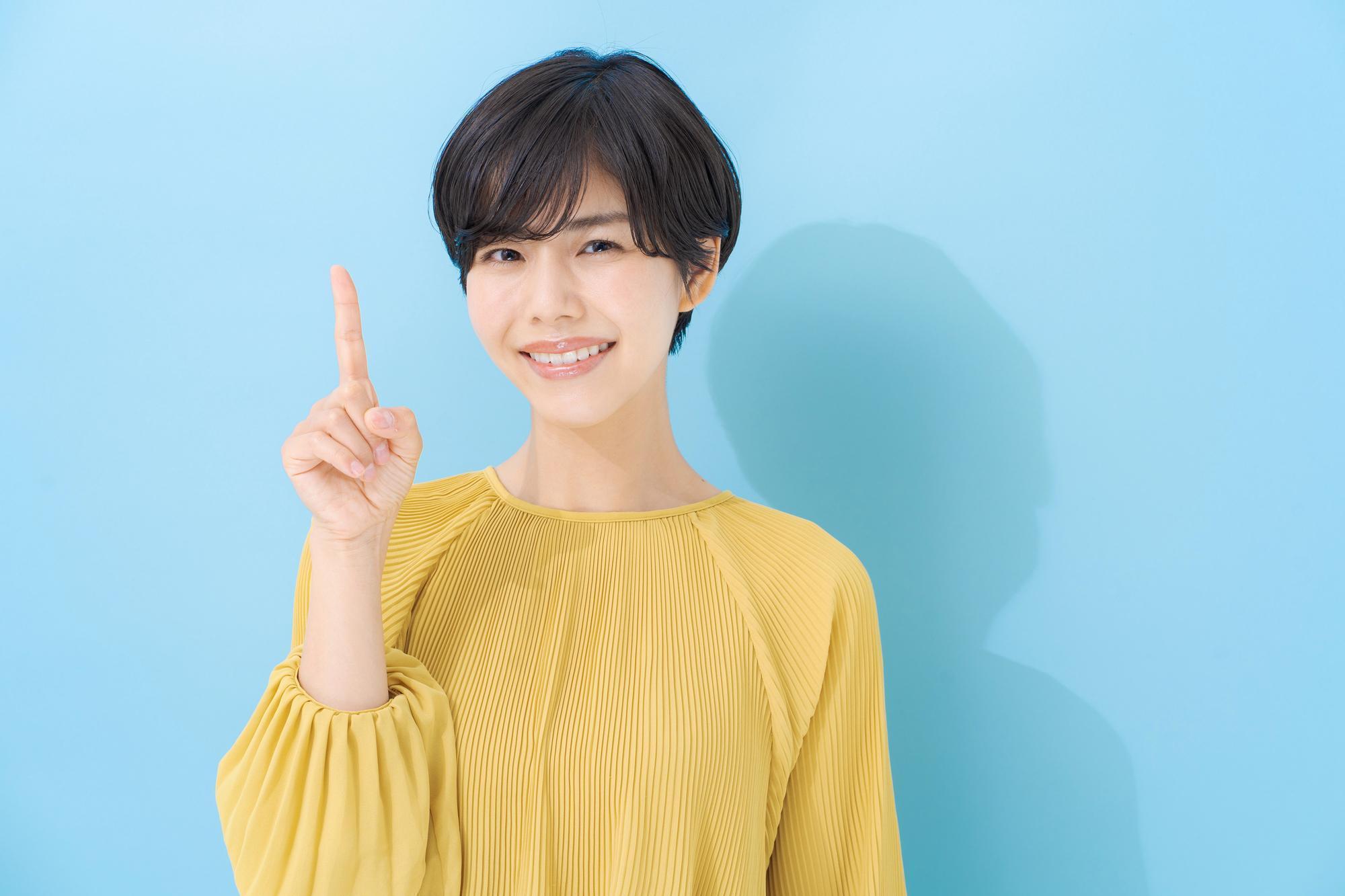 水色の背景、人さし指を立て、カメラ目線の笑顔の女性